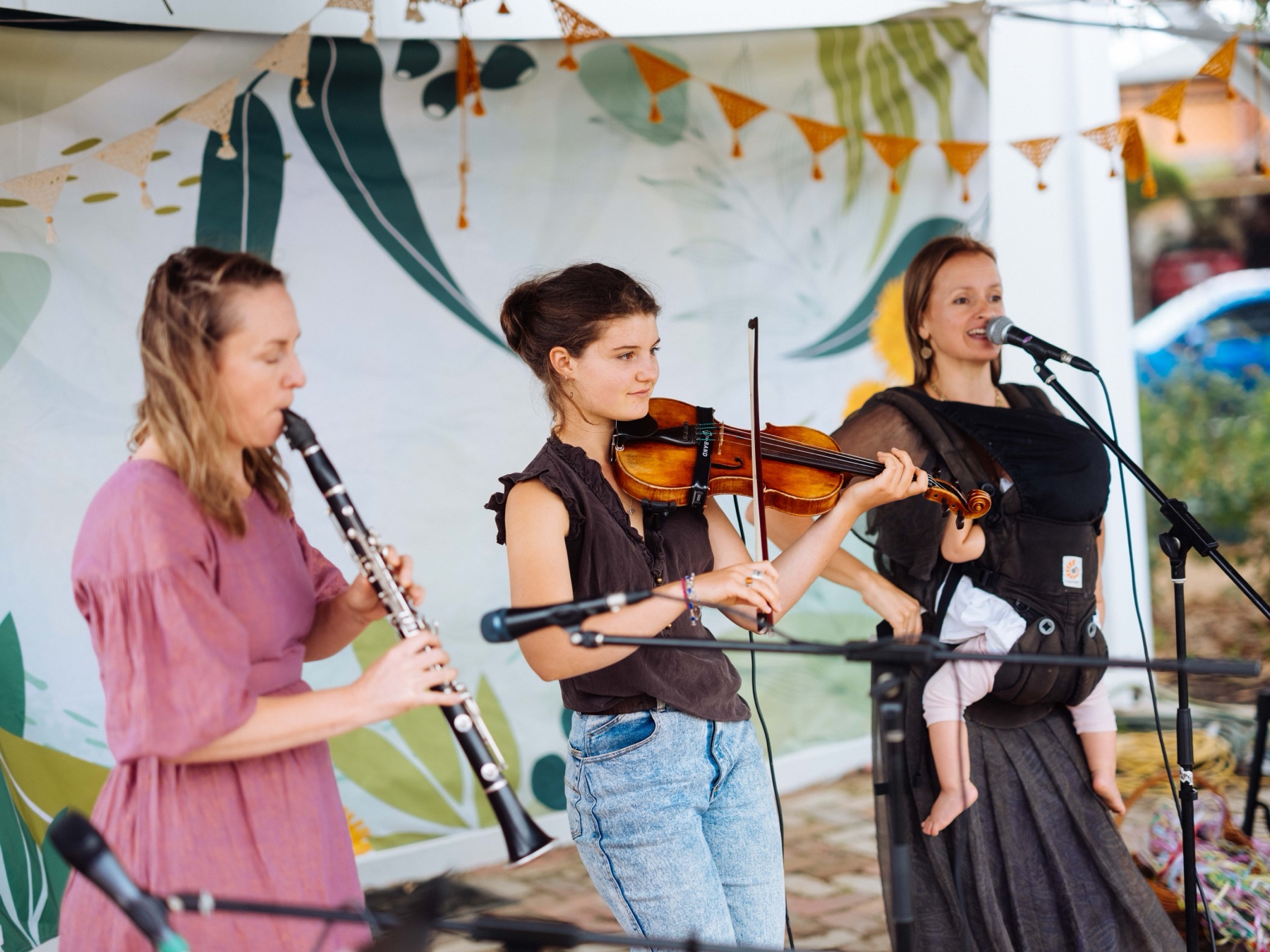 Rachel singing with a baby in a carrier alongside a clarinet and violin player