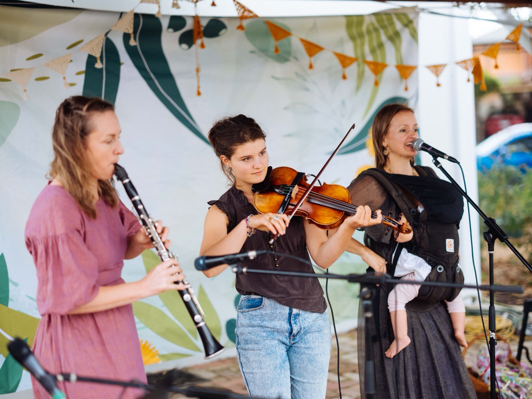 Rachel singing into a microphone with a baby in a carrier, beside a violinist and a clarinettist